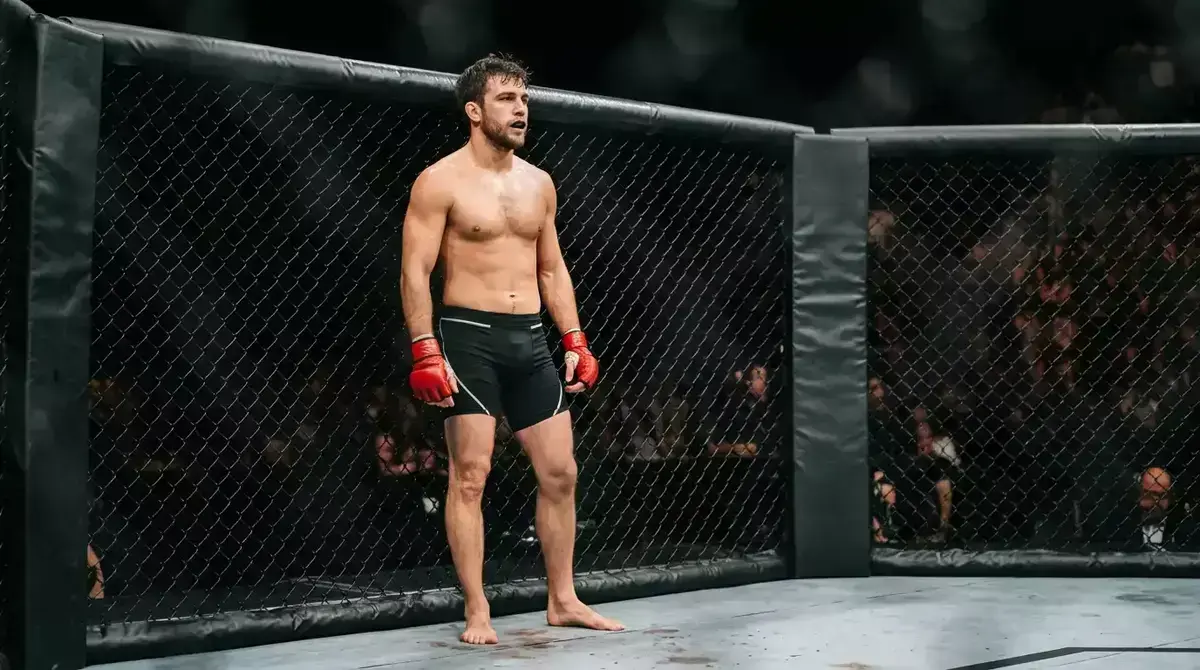 Luchador de MMA con guantes abiertos apoyado en la reja del octágono durante la instrucción del árbitro